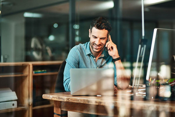 A man seated at a desk, engaged in a phone conversation while working on his laptop.
